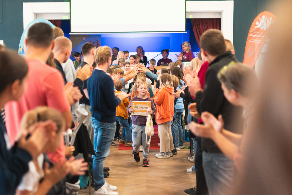 Ein Bild von dem Ende der Abschlussveranstaltung des Schüler*innenHaushalts 2024, Brandenburg. Links und rechts im Bild stehen Menschen die nach vorne in die Mitte schauen. In der Mitte befindet sich die Bühne , von dieser kommen, in diesem Augenblick viele Kinder heruntergelaufen , die kleine Geschenke und Abschlusszertifikate in der Hand halten. Die werden von den Menschen die links und rechts am Rand stehen beklatscht und beglückwünscht. © Servicestelle Jugendbeteiligung / Jonathan Gäßler 2024